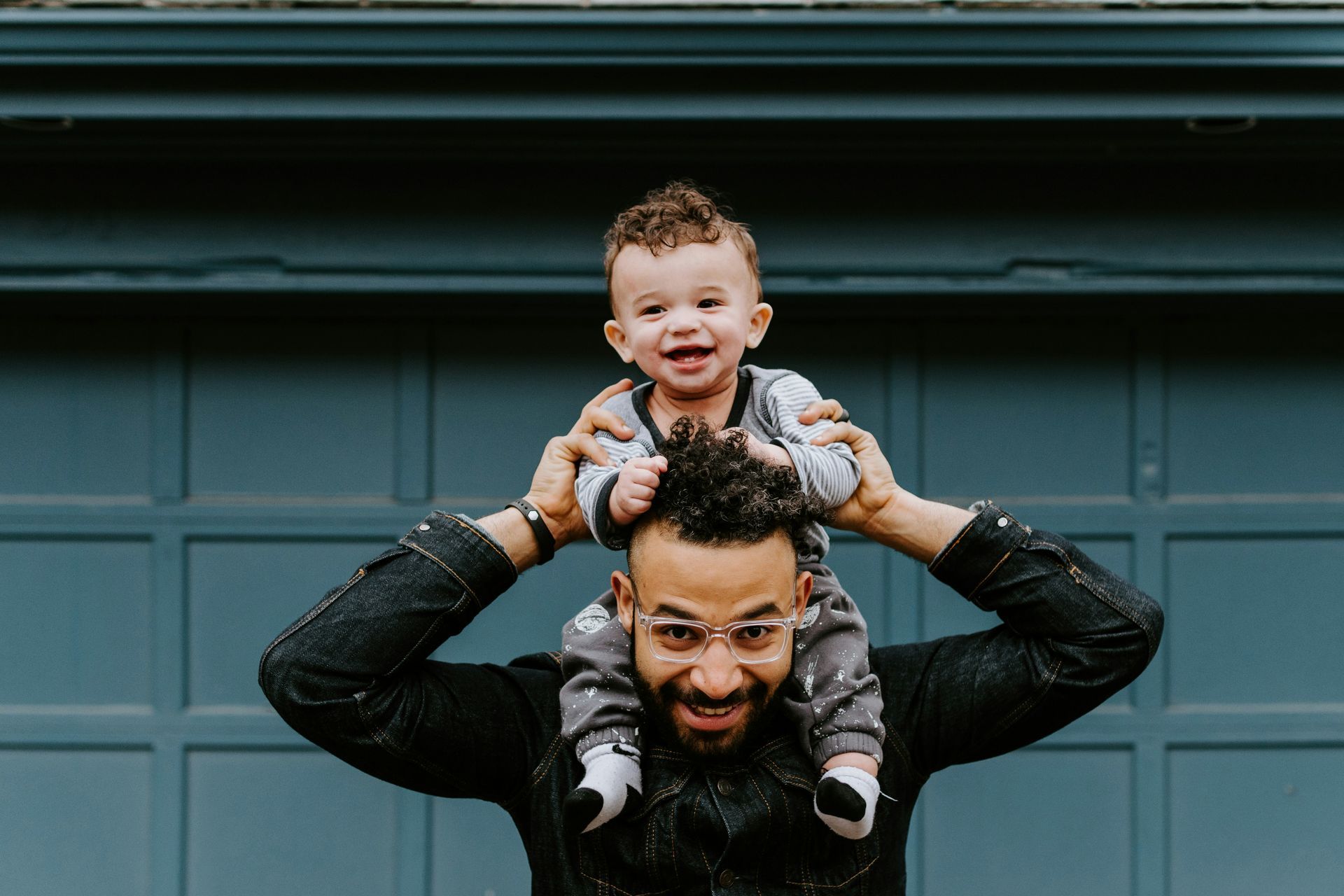 A man is carrying a baby on his shoulders in front of a garage door.